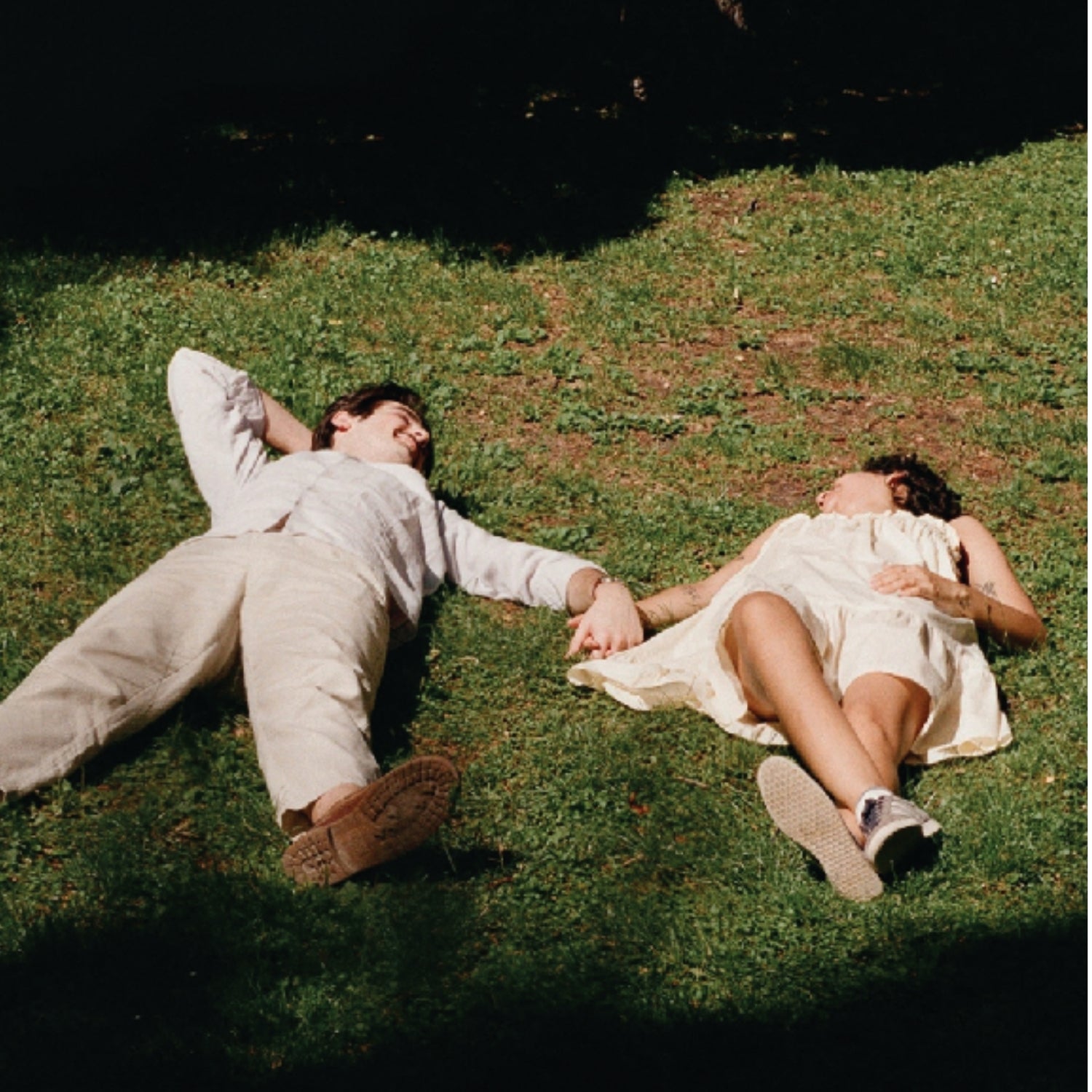 Two people lying on grass outdoors, holding hands. One wears a light beige shirt and pants, and the other wears a cream dress.