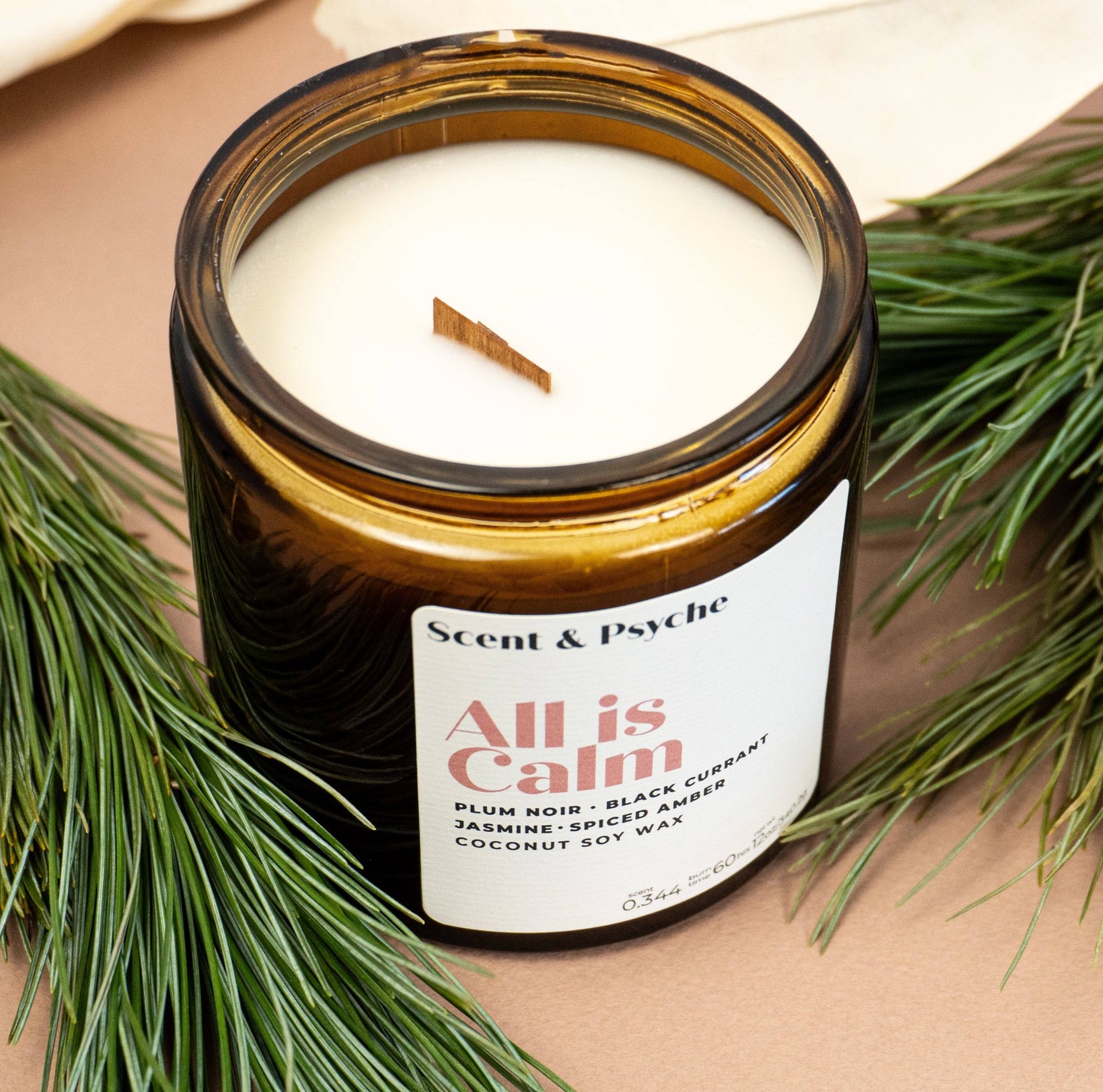 Top view of All is Calm Scented Candle in 12oz amber jar with wooden wick, surrounded by pine branches. Perfect for cozy home fragrance ambiance.