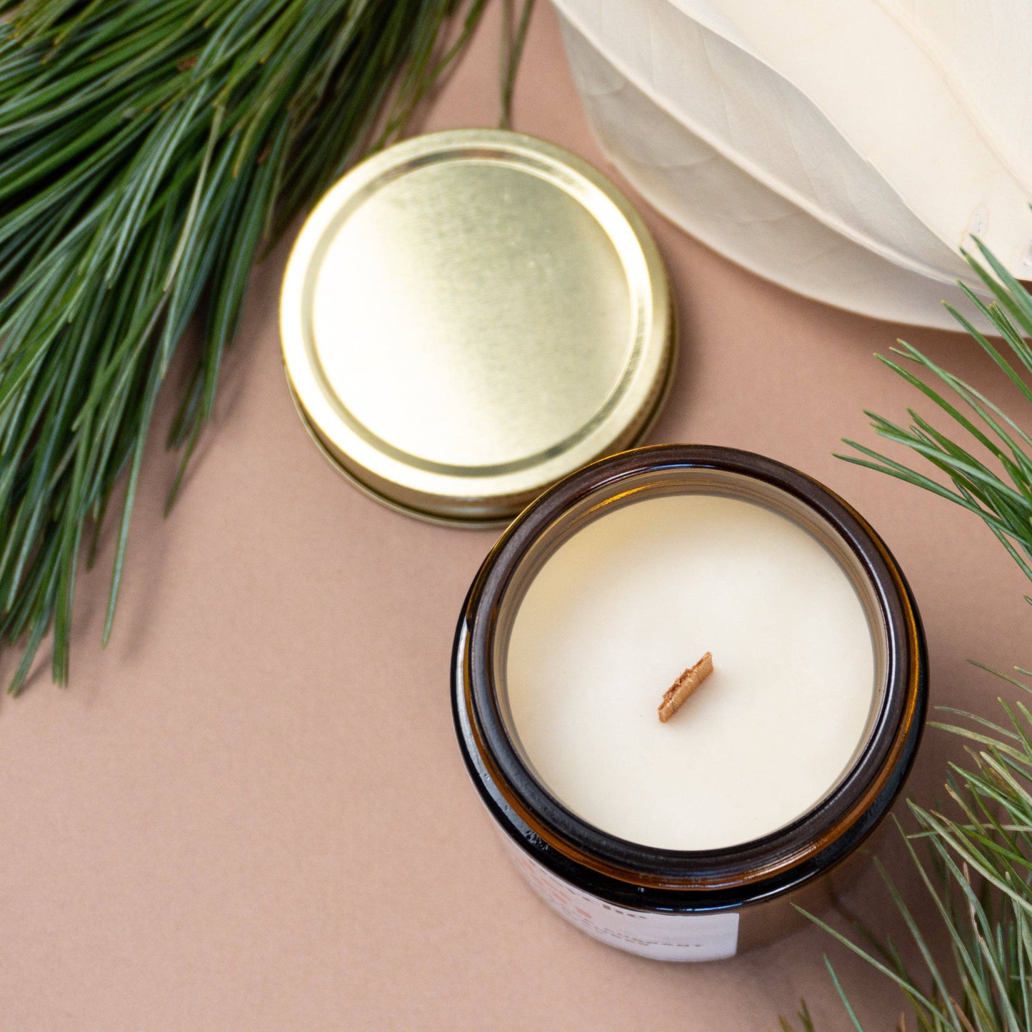 Top view of All is Calm Scented Candle in amber jar with wooden wick, gold lid off, placed among pine branches for festive ambiance.