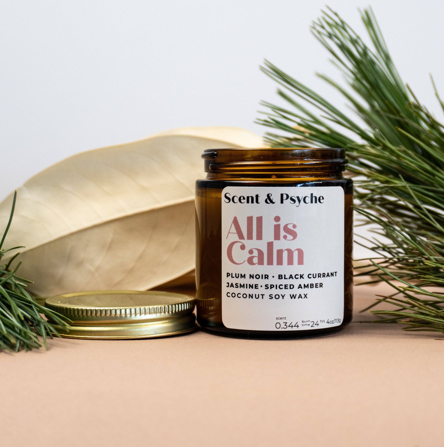 All is Calm Scented Candle in 4oz amber jar with gold lid, surrounded by pine and decorative leaves, ideal for holiday home decor.