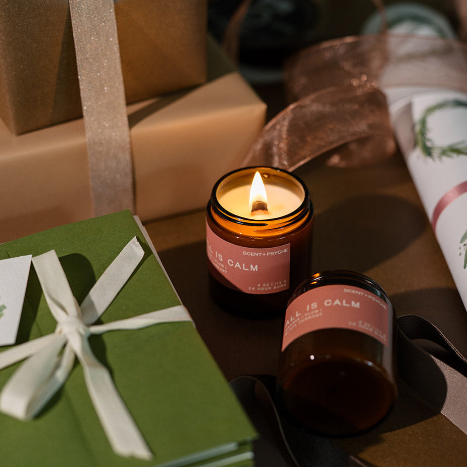 Lit All is Calm discovery candle surrounded by green-wrapped presents and rolls of wrapping paper