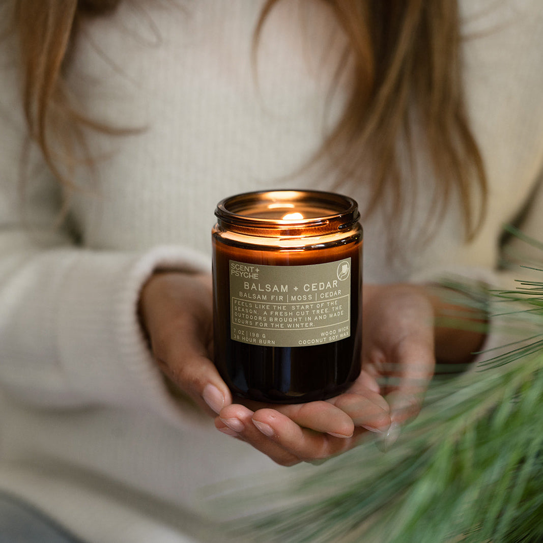 Woman in a cream sweater holding a lit Balsam + Cedar candle with pine branches in front