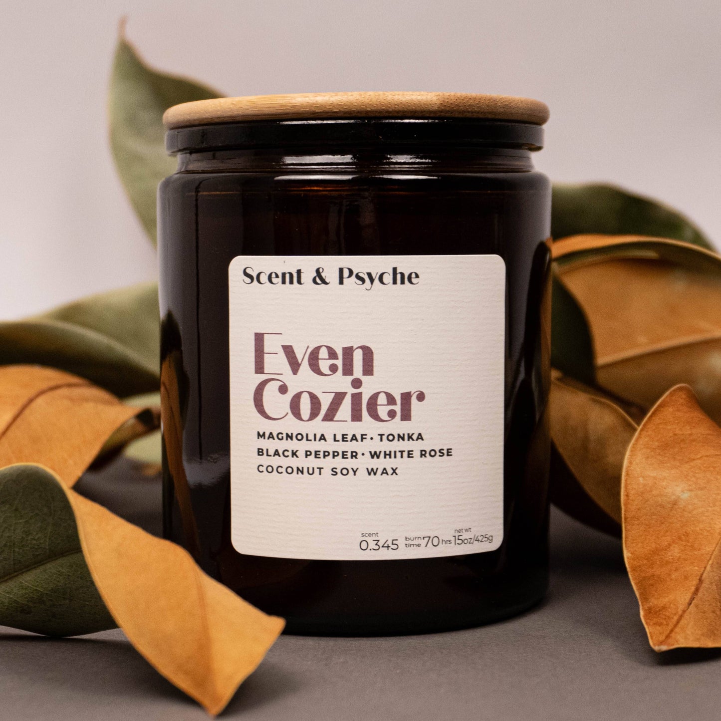 Scented Candle 15oz - Even Cozier Wood Wick Candle in amber jar with wooden lid, surrounded by dried magnolia leaves for fall decor appeal.