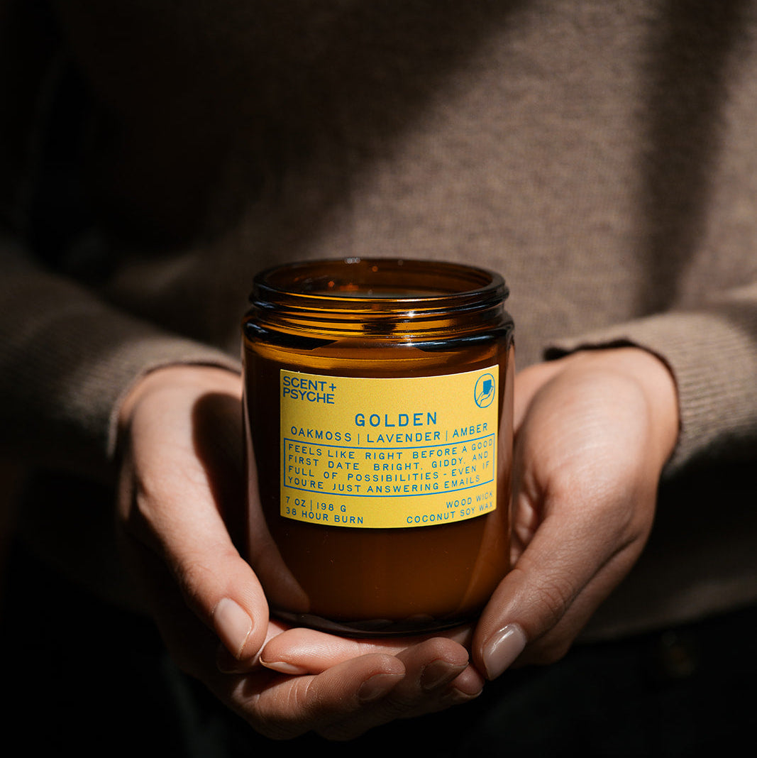 Energizing + Fresh Scented Candle Bundle | Signature Size. Golden Candle Person holding an amber jar of 'Golden' candle with a yellow label, against a dark background