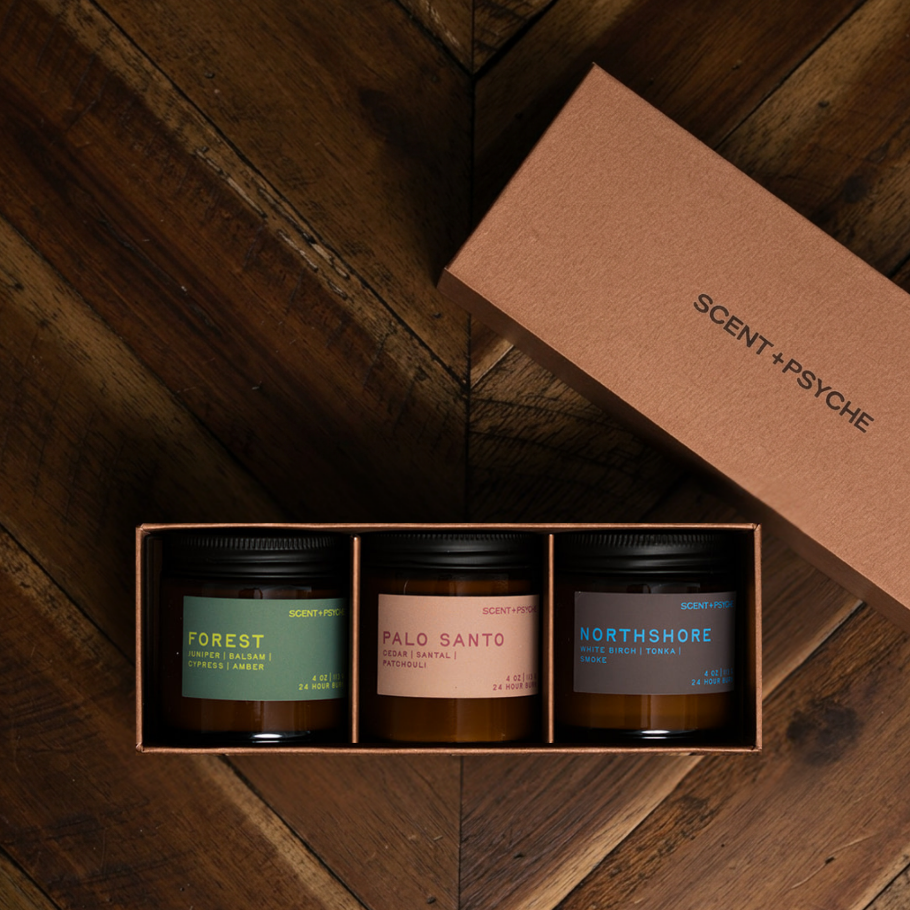 Grounding + Earthy Scented Candle Bundle | Discovery Size. A boxed set of three scented candles in glass jars with black lids, displayed in an open kraft paper box on a dark wood surface. The lid beside the box reads 'SCENT+PSYCHE'.