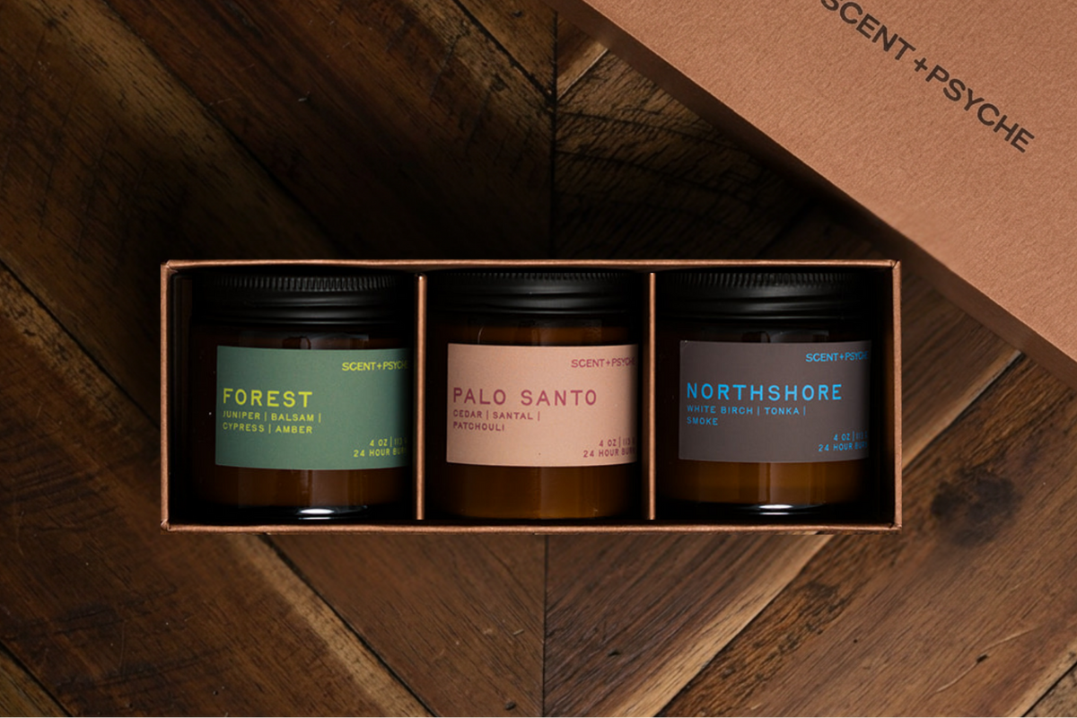 Grounding + Earthy Scented Candle Bundle | Discovery Size. A boxed set of three scented candles in glass jars with black lids, displayed in an open kraft paper box on a dark wood surface. The lid beside the box reads 'SCENT+PSYCHE'.