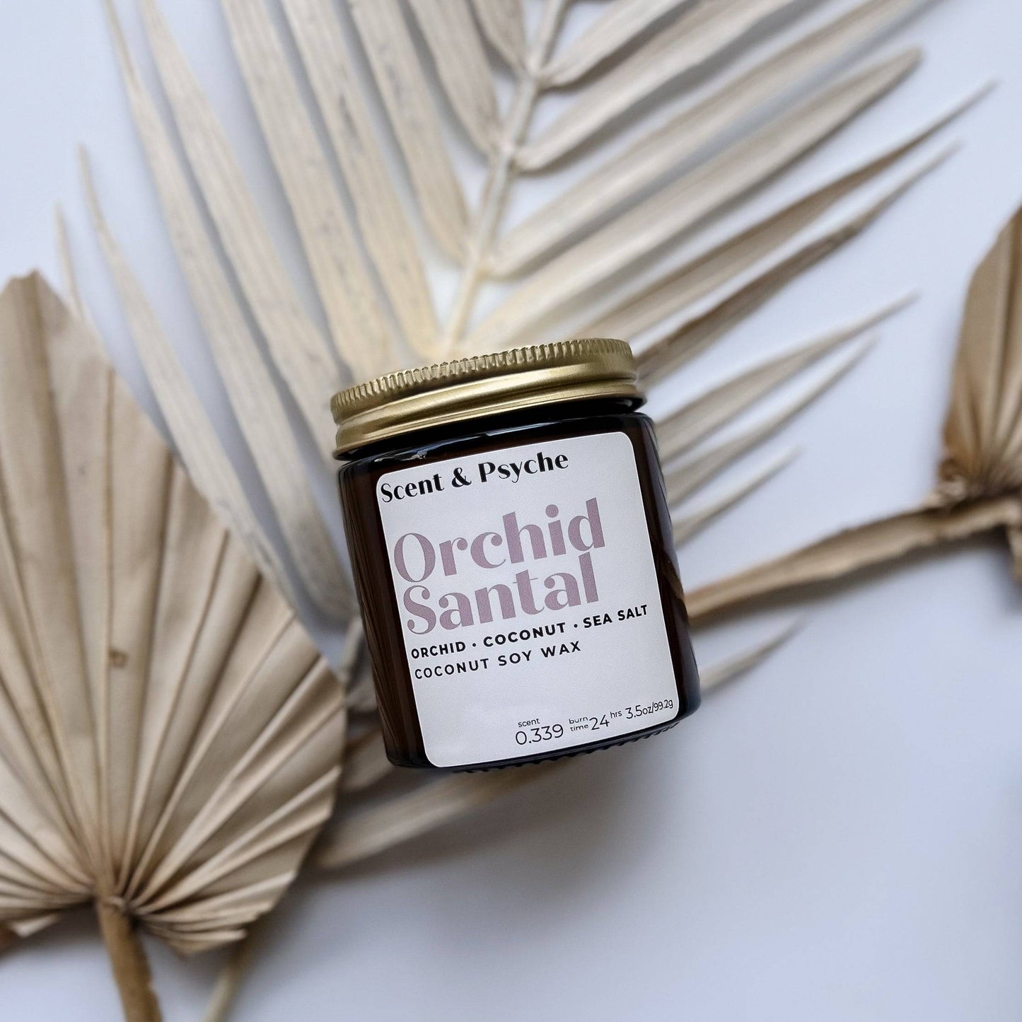 Orchid Santal Scented Candle in a 4oz amber jar with a gold lid, placed on dried palm leaves. Features a blend of orchid, coconut, and sea salt.