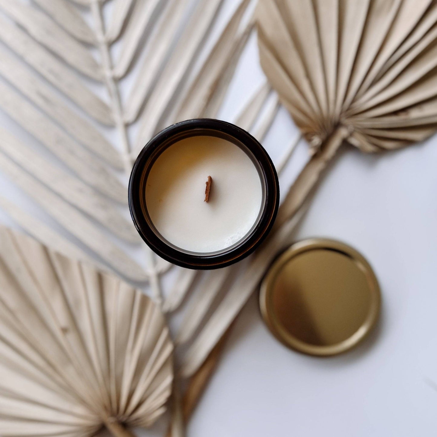 Top view of the Orchid Santal Scented Candle in a 4oz amber jar, showing a wooden wick and creamy soy wax, surrounded by dried palm leaves.