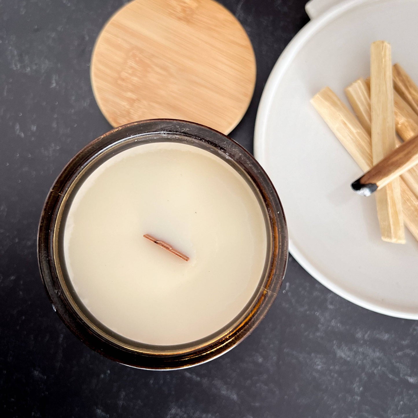 Top view of Palo Santo Scented Candle - 15oz with a wooden wick, creamy coconut soy wax, and a bamboo lid. Accompanied by Palo Santo sticks on a ceramic plate.