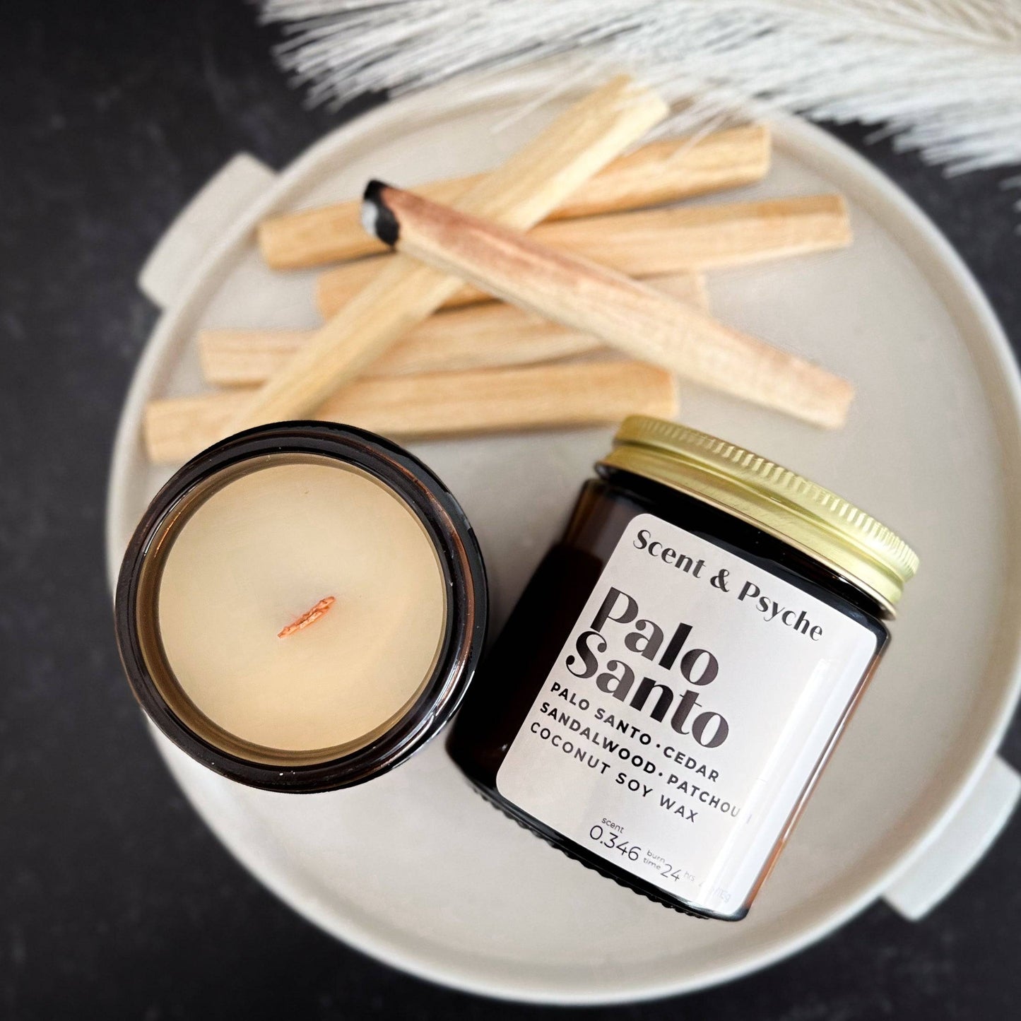 Palo Santo Scented Candle in a 4oz amber jar with a gold lid, placed on a white ceramic plate alongside Palo Santo wood sticks. The candle has a coconut soy wax blend with notes of sandalwood, cedar, and patchouli, creating an earthy and grounding aroma.