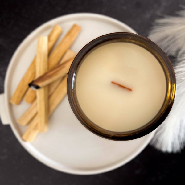 Top view of a Palo Santo Scented Candle in a 12oz amber jar with a wooden wick, placed on a tray with Palo Santo sticks. Cozy and earthy home fragrance.