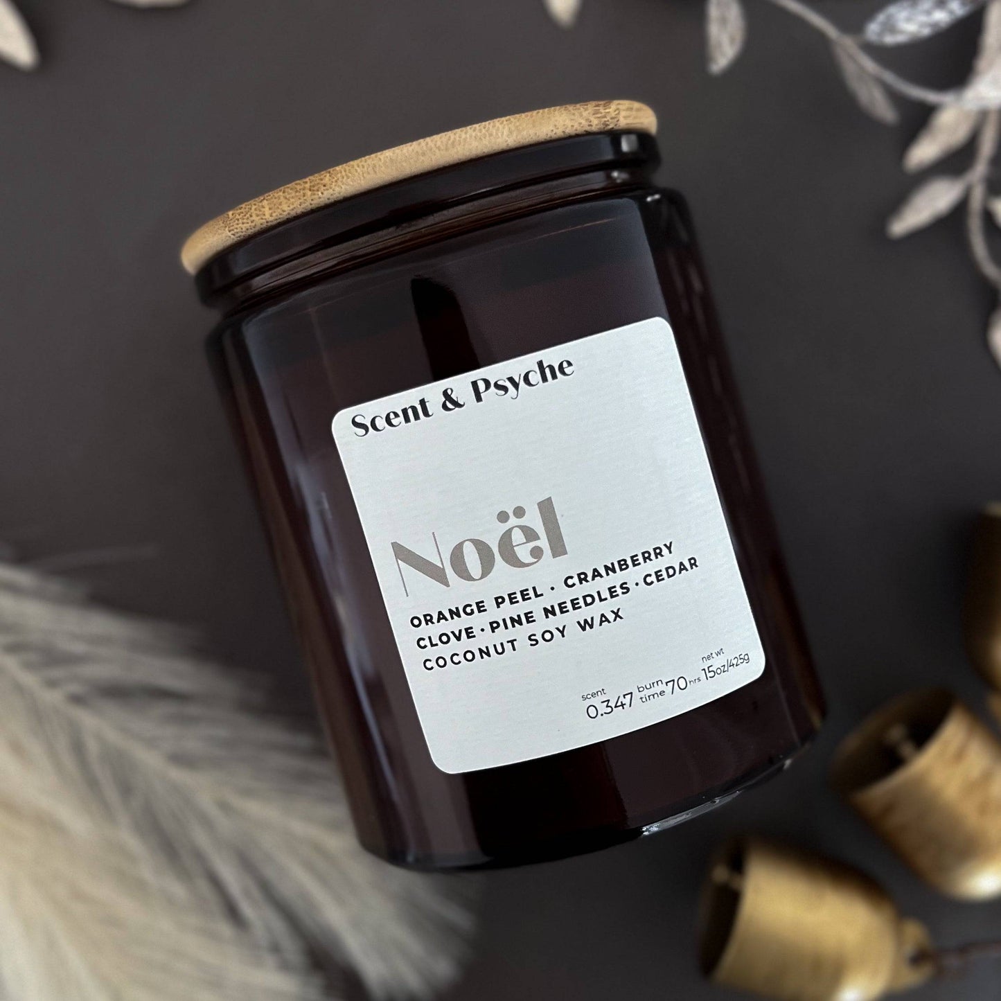 Noel Wood Wick Scented Candle in a 15 oz amber jar with a wooden lid. Features festive notes of orange peel, cranberry, clove, pine needles, and cedar.