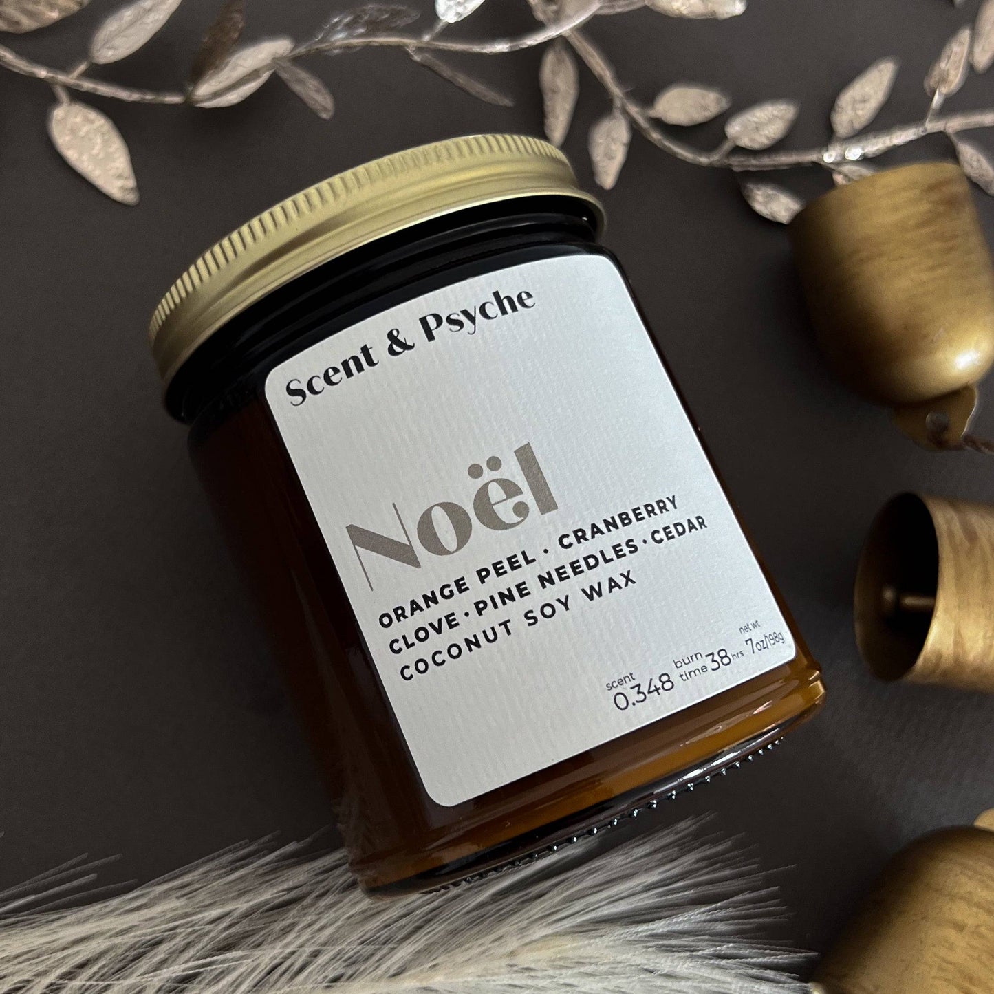 Noel Wood Wick Scented Candle in a 7oz amber jar with a gold lid. Features a festive blend of orange peel, cranberry, clove, pine needles, and cedar.