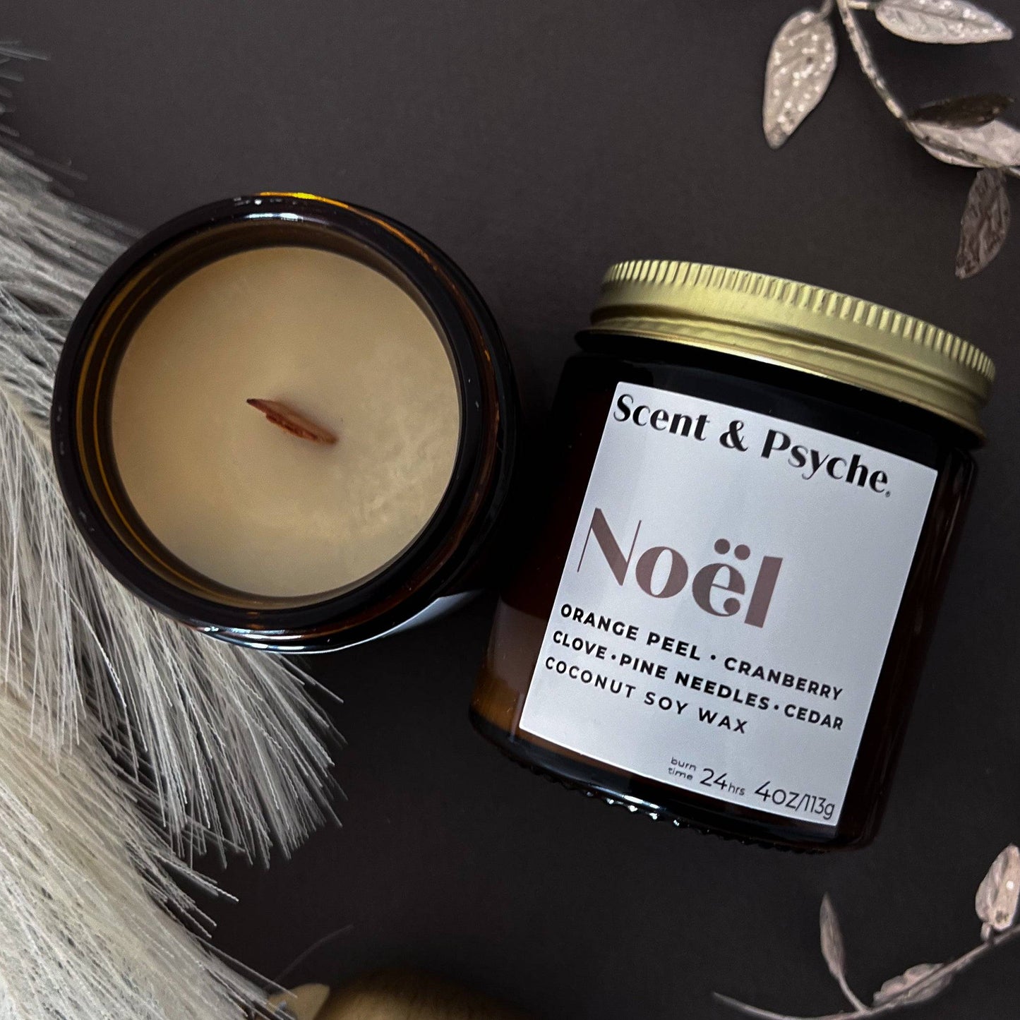Noel wood wick scented candle in a 4oz amber jar with a gold lid. Features a coconut soy wax blend with notes of orange peel, cranberry, clove, pine needles, and cedar.
