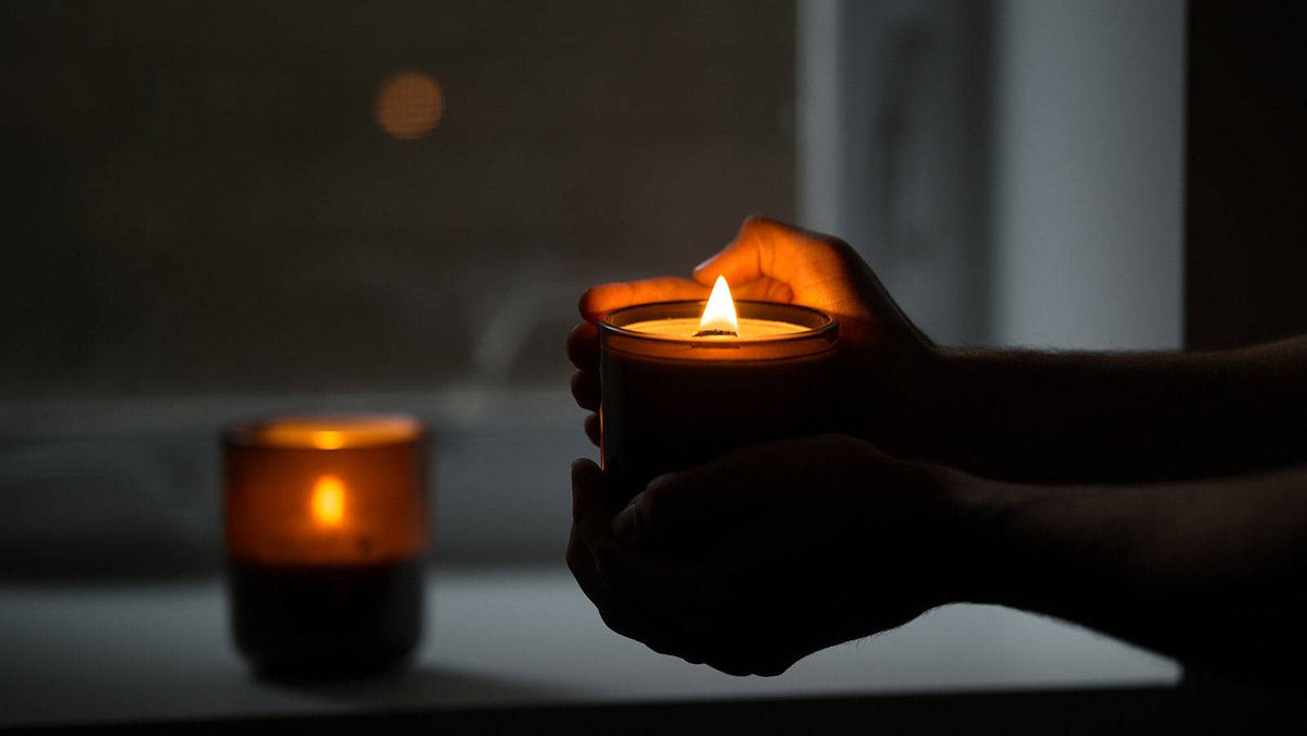 Hands holding a lit amber glass wood wick candle jar in a dimly lit room, with a second candle blurred in the background near a window.