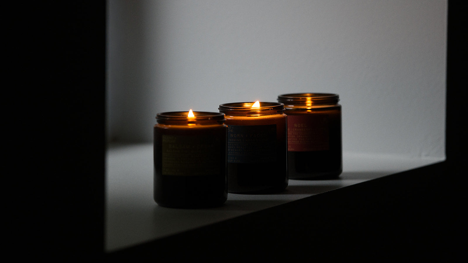 Three lit wood wick candles in dark amber glass jars sit on a white ledge against a softly lit gray wall.