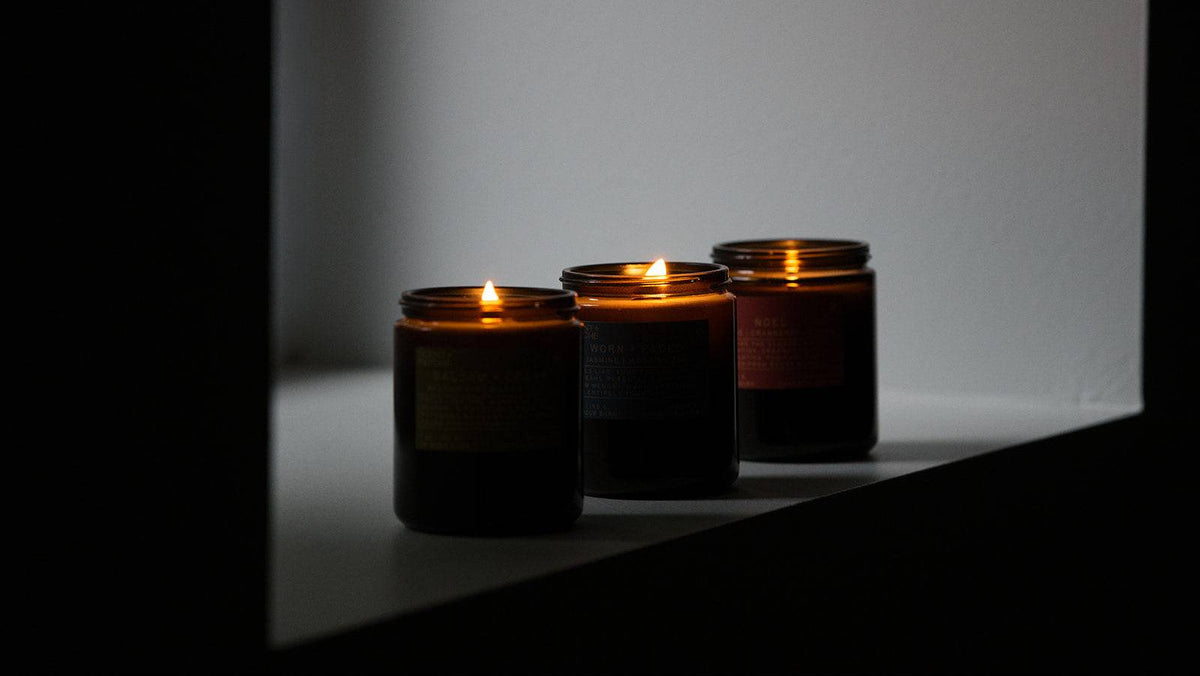 Three lit wood wick candles in dark amber glass jars sit on a white ledge against a softly lit gray wall.