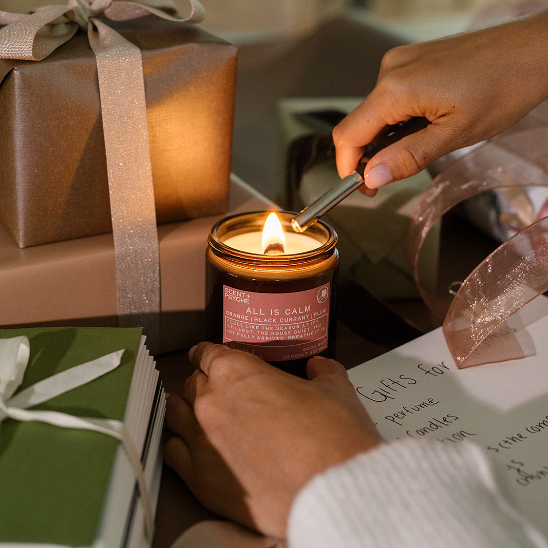 Lit All is Calm candle beside hands wrapping a holiday gift in green paper