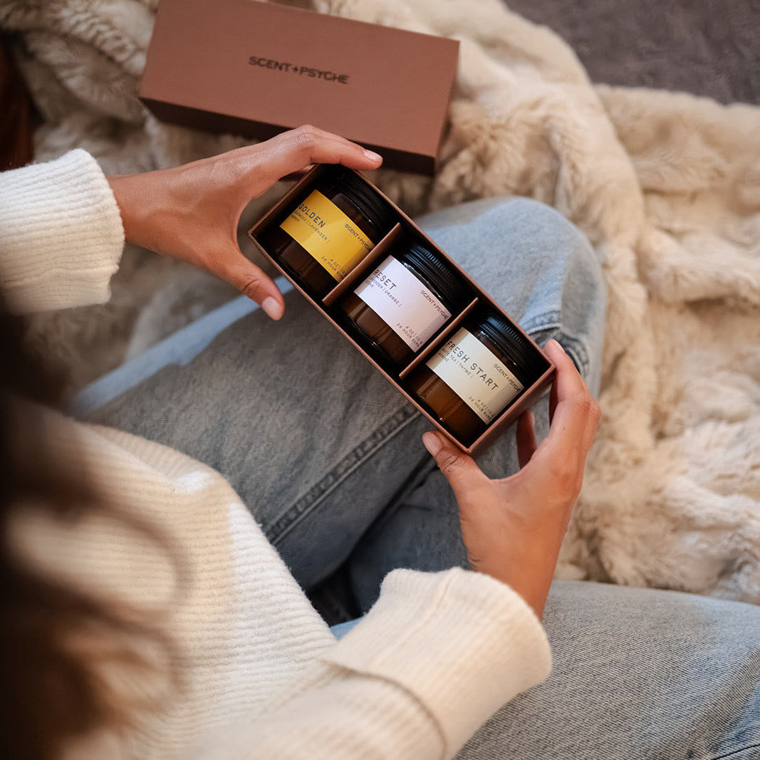 Scent + Psyche Energizing + Fresh soy candle discovery bundle in custom gift box. A person in a cream knit sweater and light blue jeans holds an open box containing three candles from a Scent and psyche set, with a brown branded box and a beige throw blanket in the background.