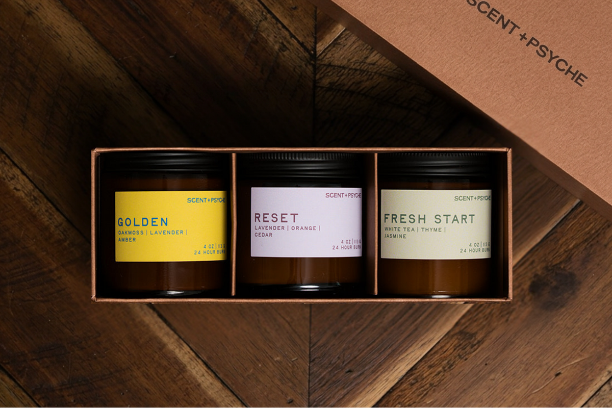 Scent + Psyche Energizing + Fresh soy candle discovery bundle in custom gift box. A three-scent candle gift set by Scent + Psyche in a brown elevated gift box, displayed on a dark wood background with the matching lid leaning against it. The candles have black lids and labels in yellow, lavender, and sage green.