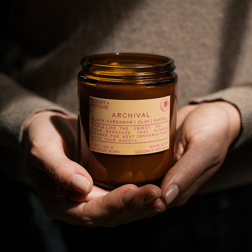 A person holds an amber glass jar candle with a cream label reading 'ARCHIVAL' in a dim, warmly lit setting.