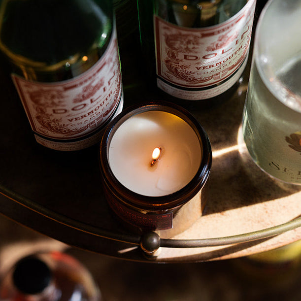 A two-tiered brass bar cart filled with assorted liquor and spirit bottles, with a small lit candle on the top tier, photographed in warm, dramatic lighting.