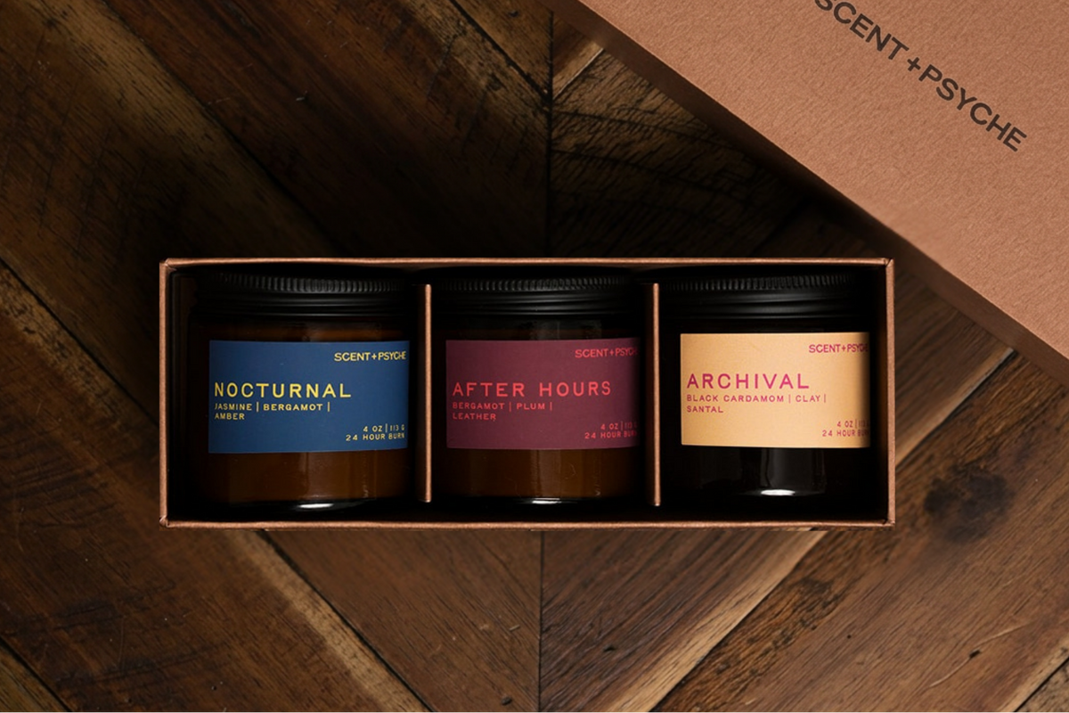 Warm + Floral Scented Candle Bundle | Discovery Size. A three-scented candle gift set by Scent+Psyche in a brown cardboard tray box, displayed on a dark wood chevron floor with the matching lid resting beside it. The candles feature dark amber glass jars with black lids and labels in navy blue, dusty rose, and warm tan.