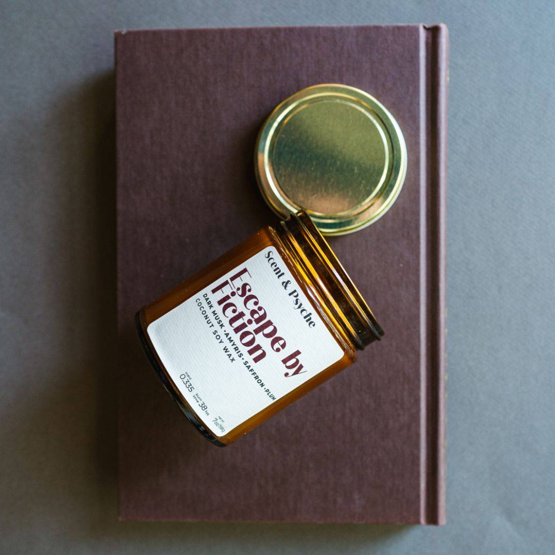 Escape by Fiction Scented Candle in a 7oz amber jar with a gold lid, placed on a brown book. A cozy home fragrance for relaxation and ambiance.
