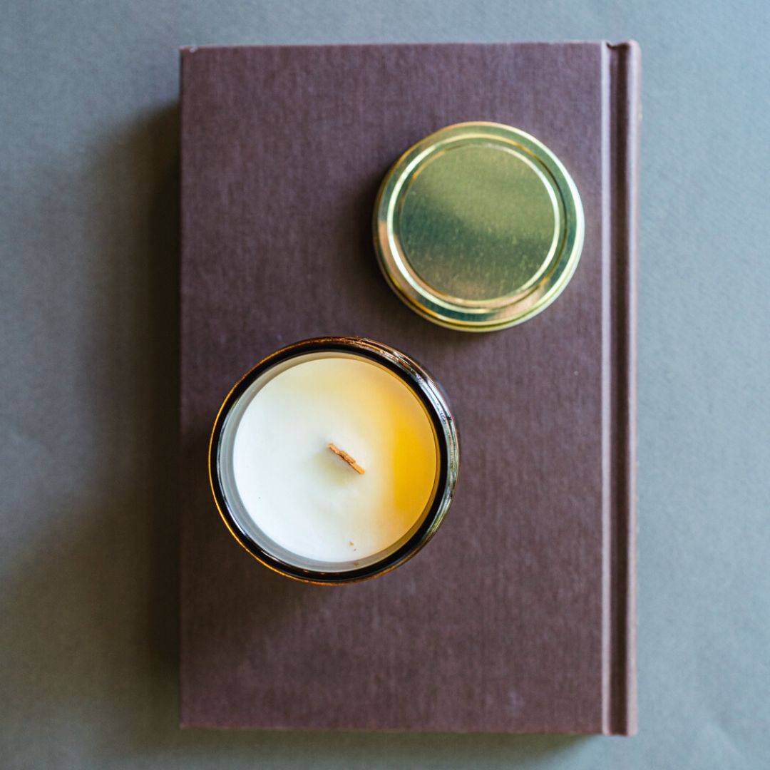 Top view of Escape by Fiction Scented Candle in a 7oz amber jar with a wooden wick, placed on a brown book. A warm and inviting home fragrance.