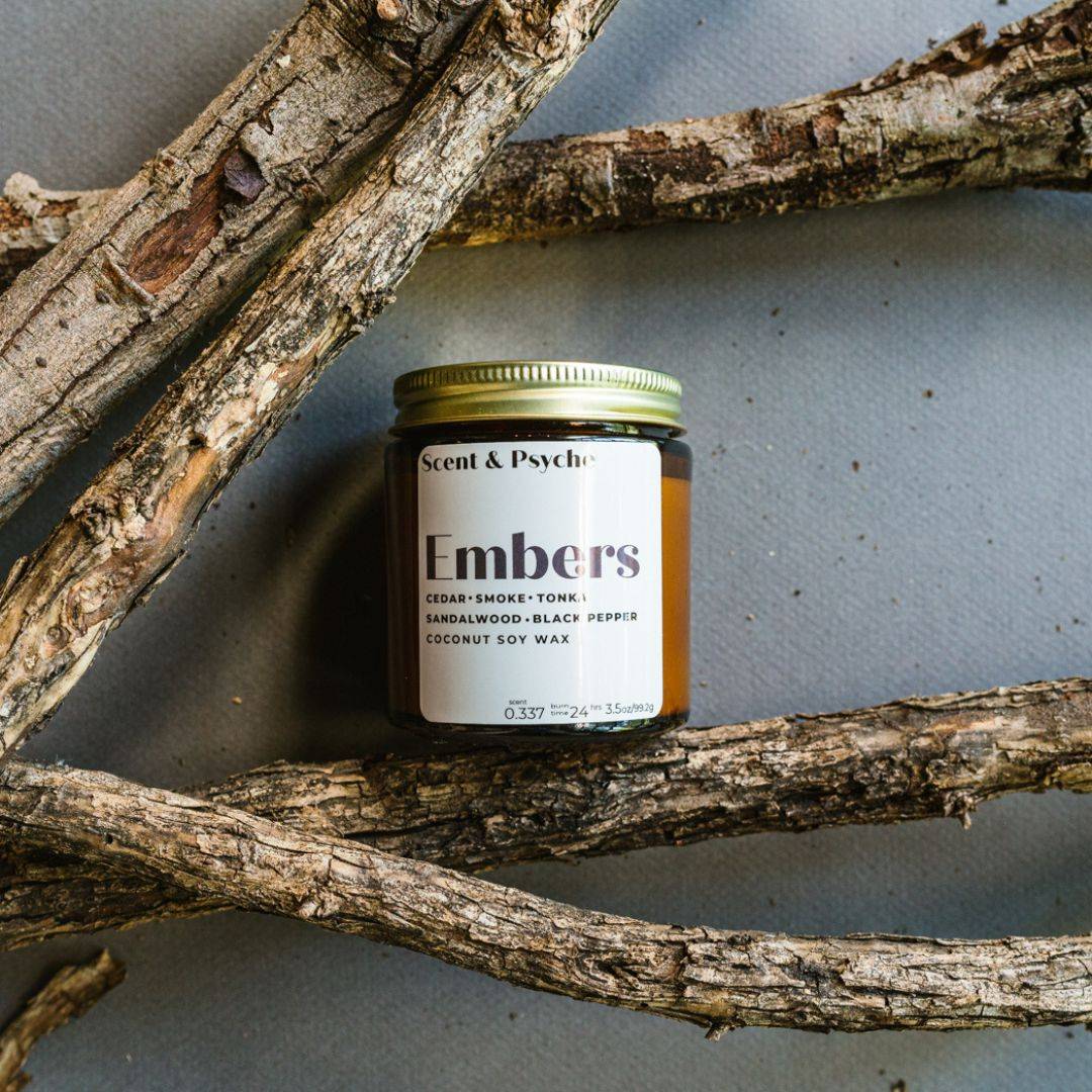 Embers Scented Candle in a 4oz amber jar with a gold lid, placed among rustic wooden branches. Features notes of cedar, smoke, and black pepper.