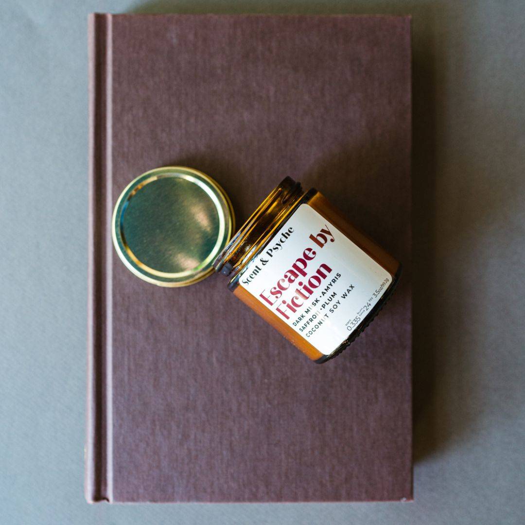 Escape by Fiction Scented Candle in a 4oz amber jar with a gold lid, placed on a brown hardcover book. A cozy home fragrance for relaxation.