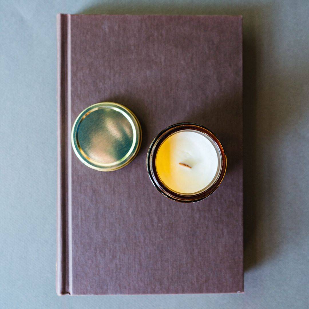 Top view of Escape by Fiction Scented Candle in a 4oz amber jar with a wooden wick, placed on a brown book. A stylish home decor fragrance piece.