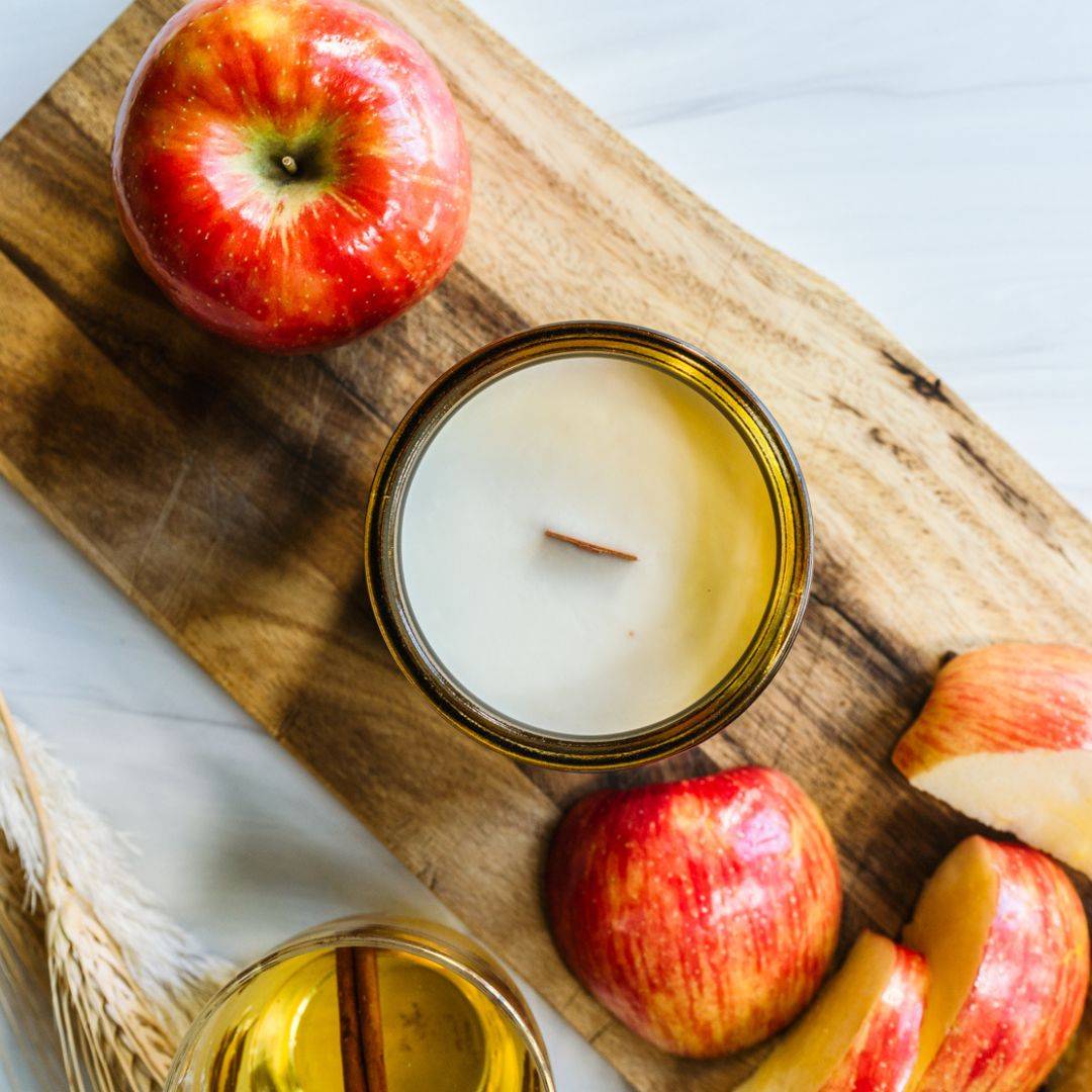Top view of Cider House 12oz amber jar scented candle with a wooden wick, surrounded by fresh apples. Cozy fall fragrance for home and garden decor.