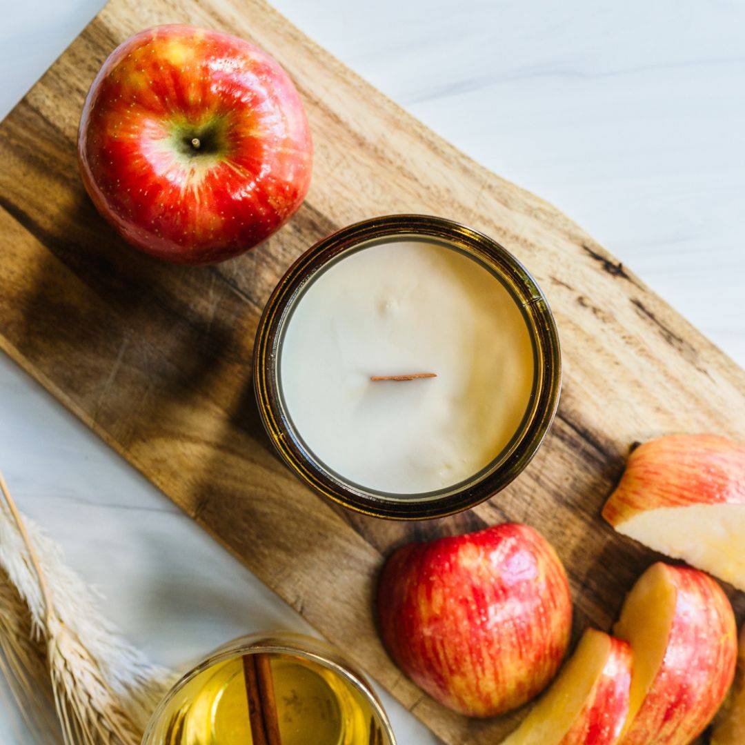 Top view of Cider House 15oz amber jar scented candle with a wooden wick, surrounded by fresh apples on a rustic wooden board. Ideal for autumn vibes.