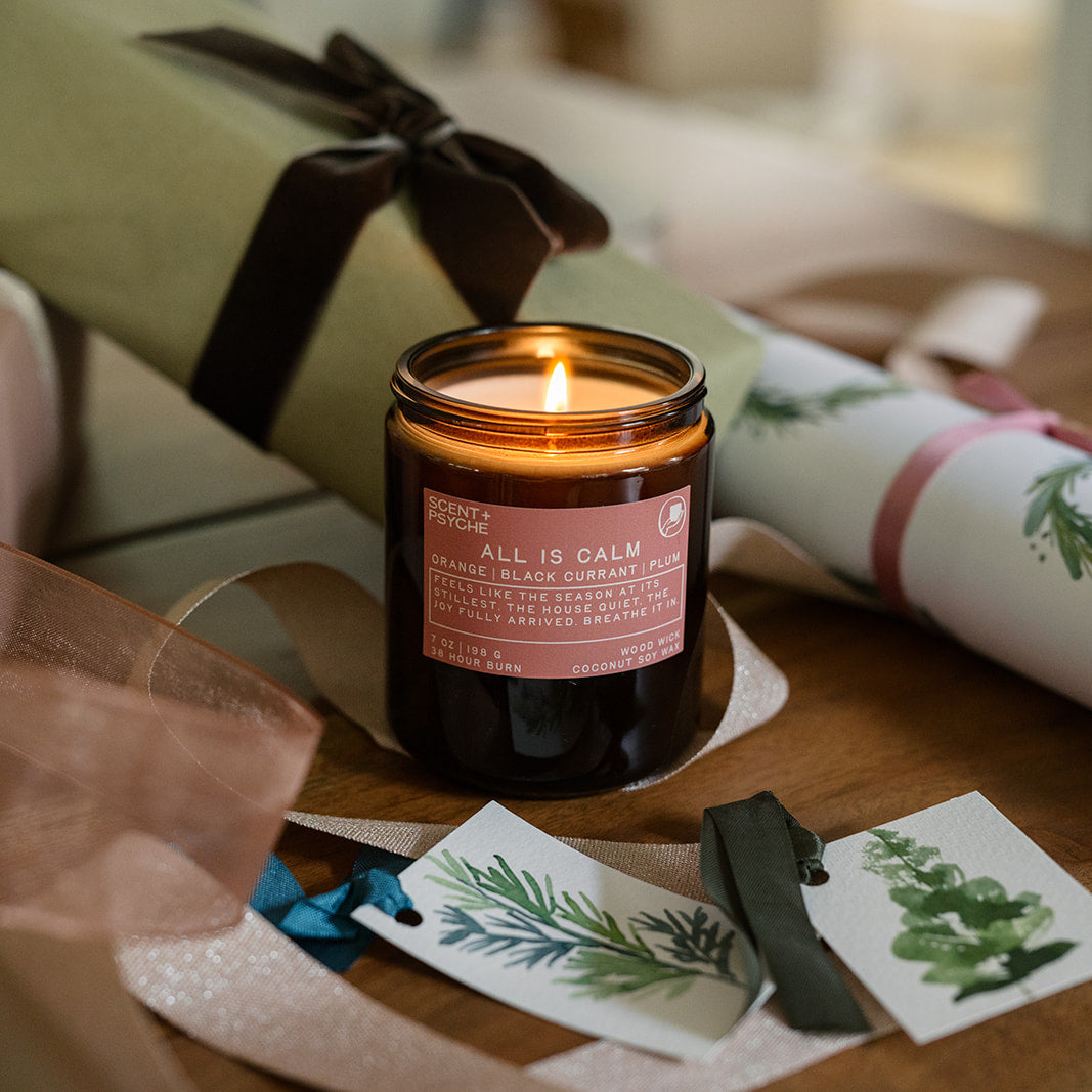 Lit All is Calm candle among wrapped holiday gifts and gift tags