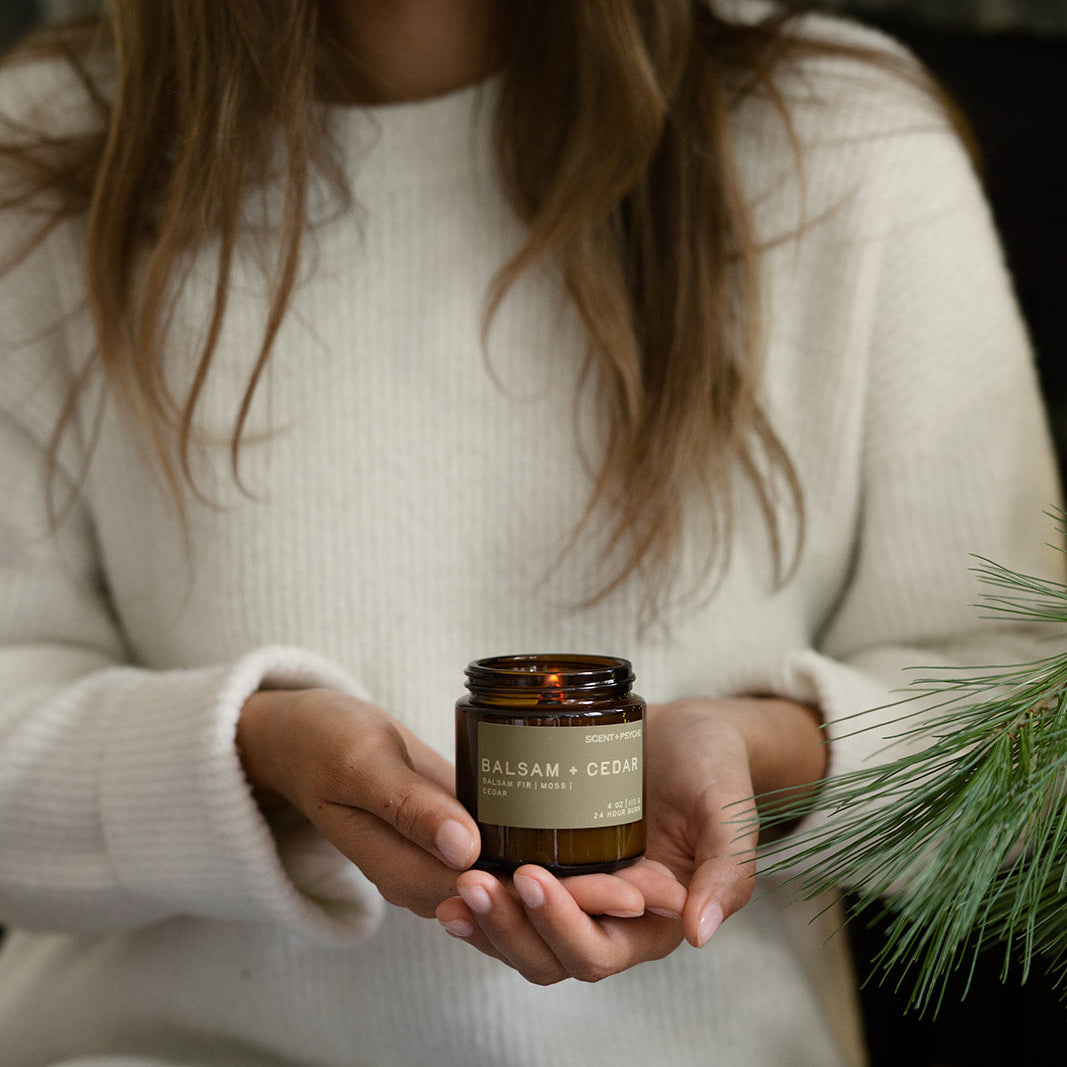 Woman in a white knit sweater holding a Balsam + Cedar candle behind pine branches