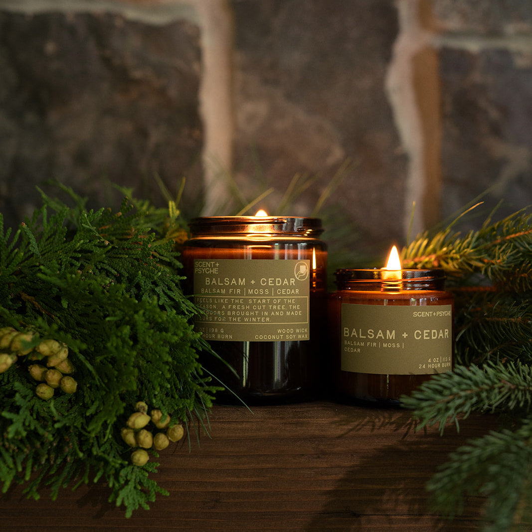 Two lit Balsam + Cedar candles with evergreen branches and pinecones against a stone wall