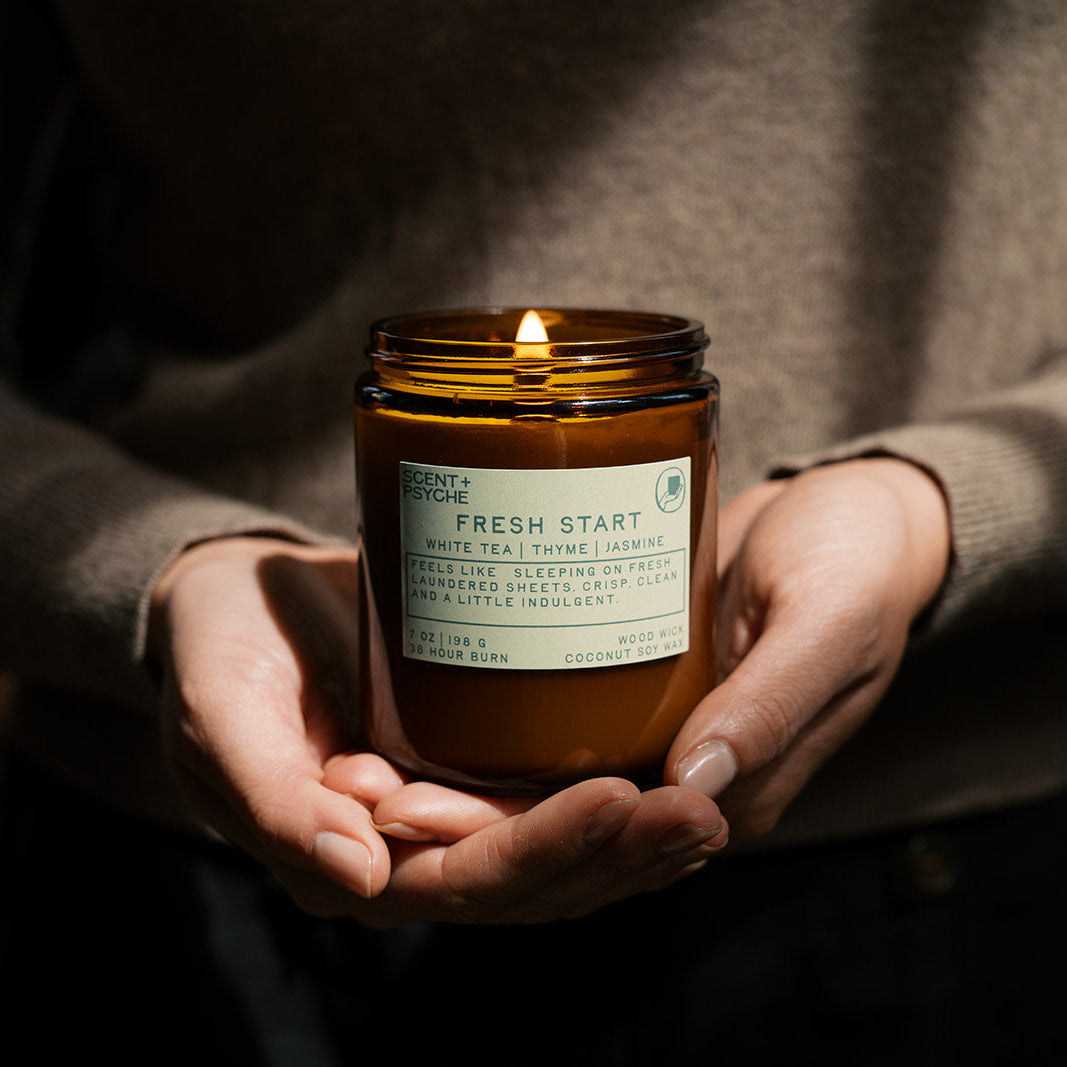 Person holding a lit candle in a jar labeled 'Fresh Start' against a dark background