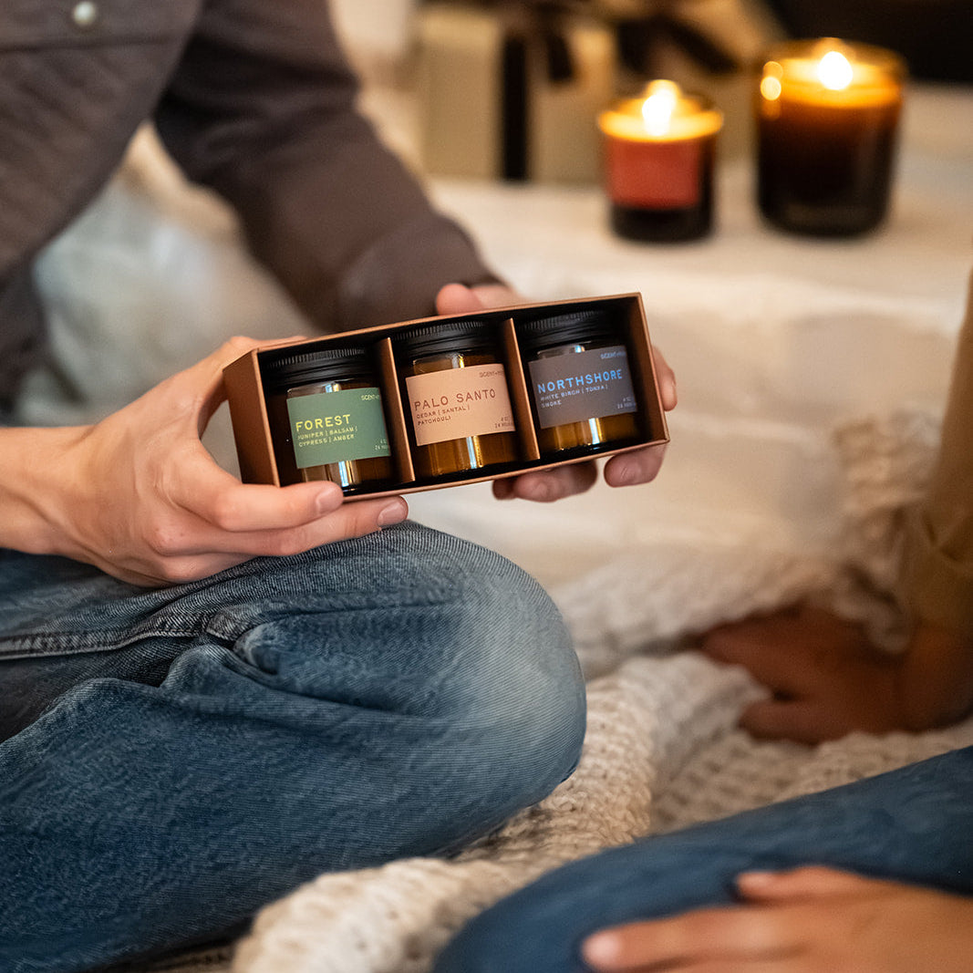 Grounding + Earthy Scented Candle Bundle | Discovery Size A person holds a small gift box with three scented candles in a cozy indoor setting, with a fireplace, wrapped gifts, and lit candles in the background.