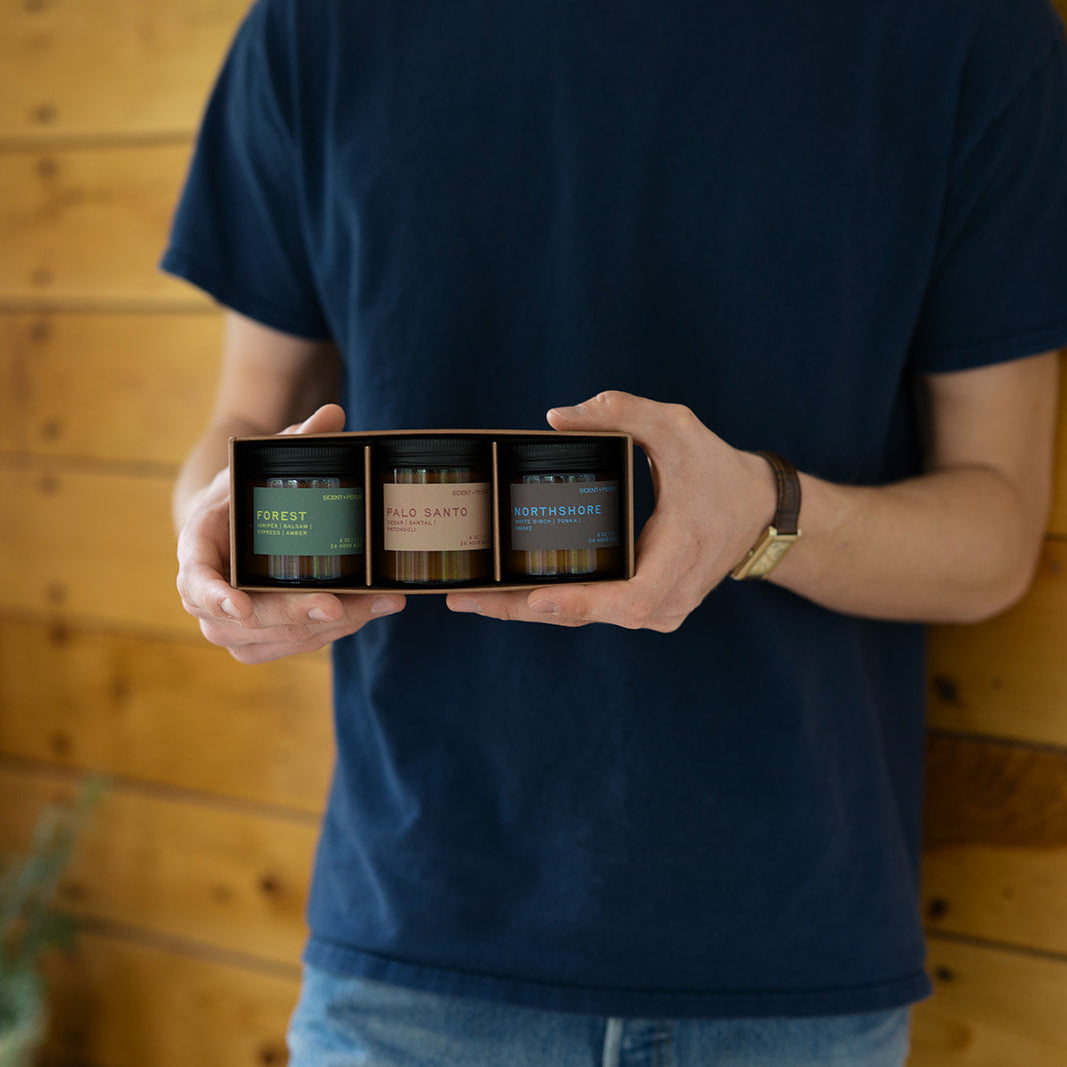 Grounding + Earthy Scented Candle Bundle | Discovery Size.  A person in a navy blue t-shirt holds a wooden gift box containing three small jars with green, brown, and blue labels, set against a rustic wood-paneled wall.