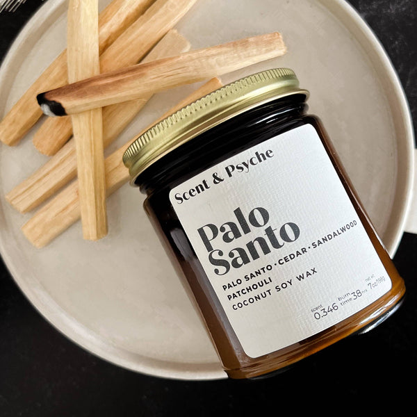 Palo Santo Scented Candle in a 7oz amber jar with a gold lid, featuring a minimalist label. Placed on a ceramic plate with Palo Santo wood sticks.