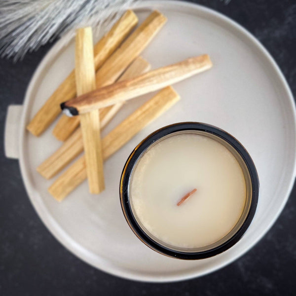 Top view of an unlit Palo Santo Scented Candle in a 7oz amber jar with a wooden wick. Displayed on a ceramic plate with Palo Santo wood sticks.