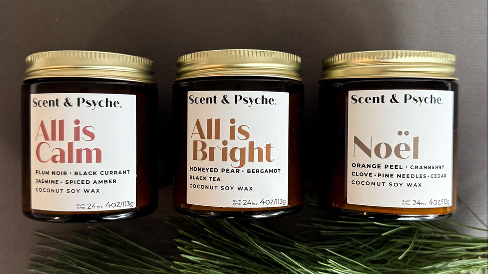 Three candles labeled 'All is Calm', 'All is Bright', and 'Noél' by Scent & Psyche on a dark background with greenery.
