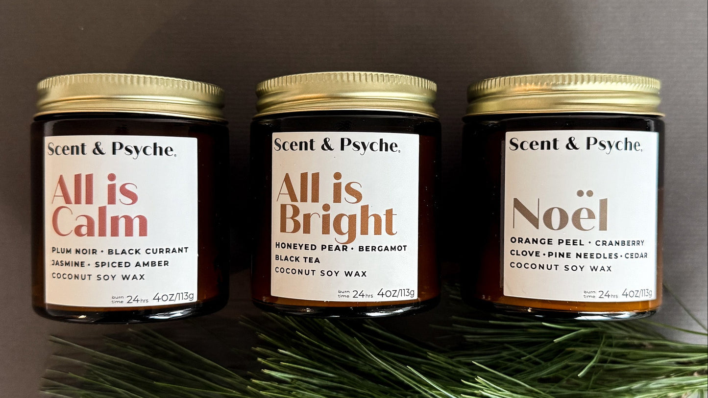Three candles labeled 'All is Calm', 'All is Bright', and 'Noél' by Scent & Psyche on a dark background with greenery.