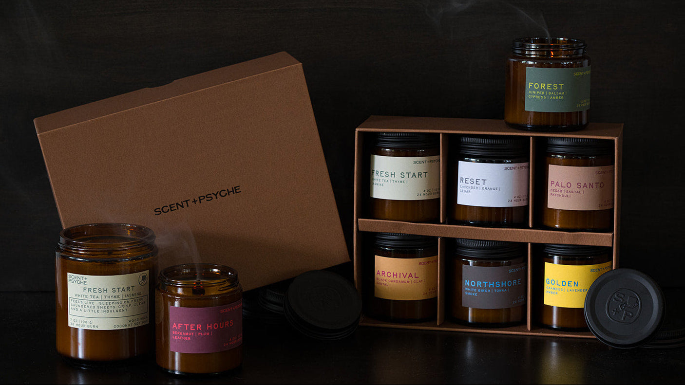 A candle gift set from Scent-Psychoe on a dark wooden surface against a dark wood background. The set includes a brown cardboard gift box, a wooden 2x3 tray holding six amber glass candles with various labels
