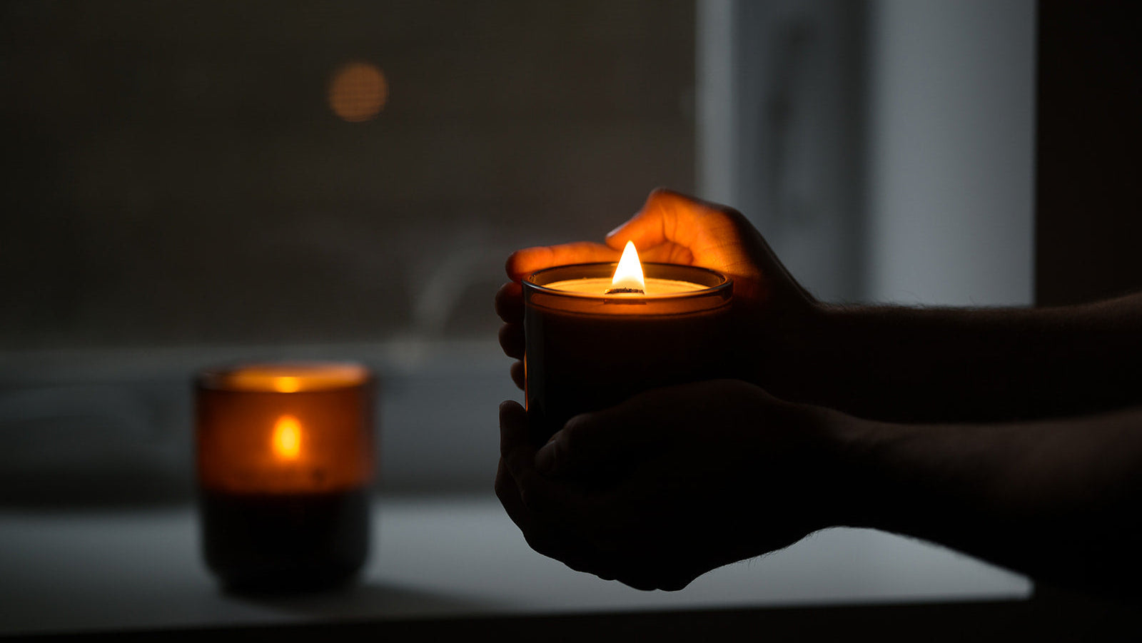 Hands holding a lit amber glass wood wick candle jar in a dimly lit room, with a second candle blurred in the background near a window.