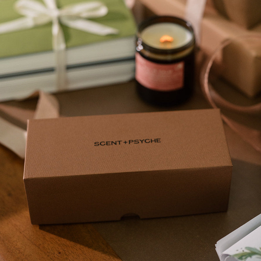 A Scent + Psyche candle in a gift box are displayed on a wooden table alongside other wrapped gift boxes and a green notebook.