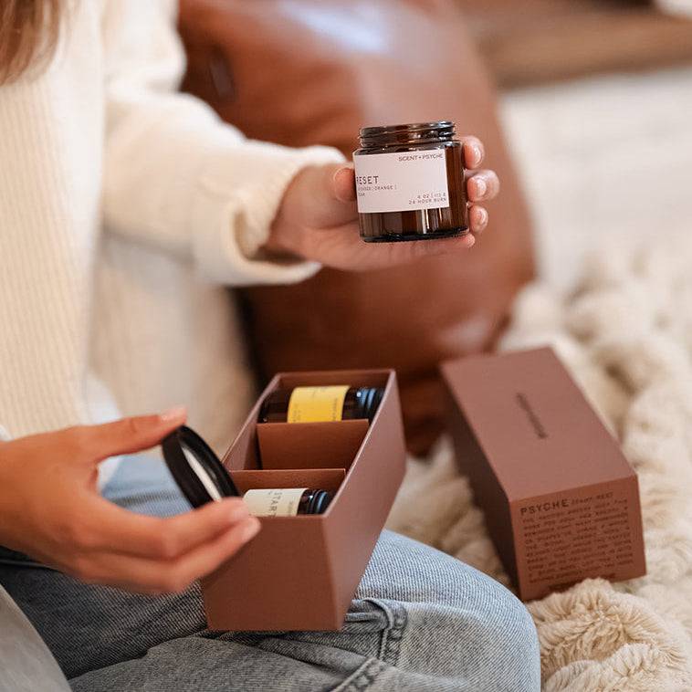 A woman sitting on a cozy rug in front of a fireplace, holding an amber glass jar and an open brown gift box with matching packaging.
