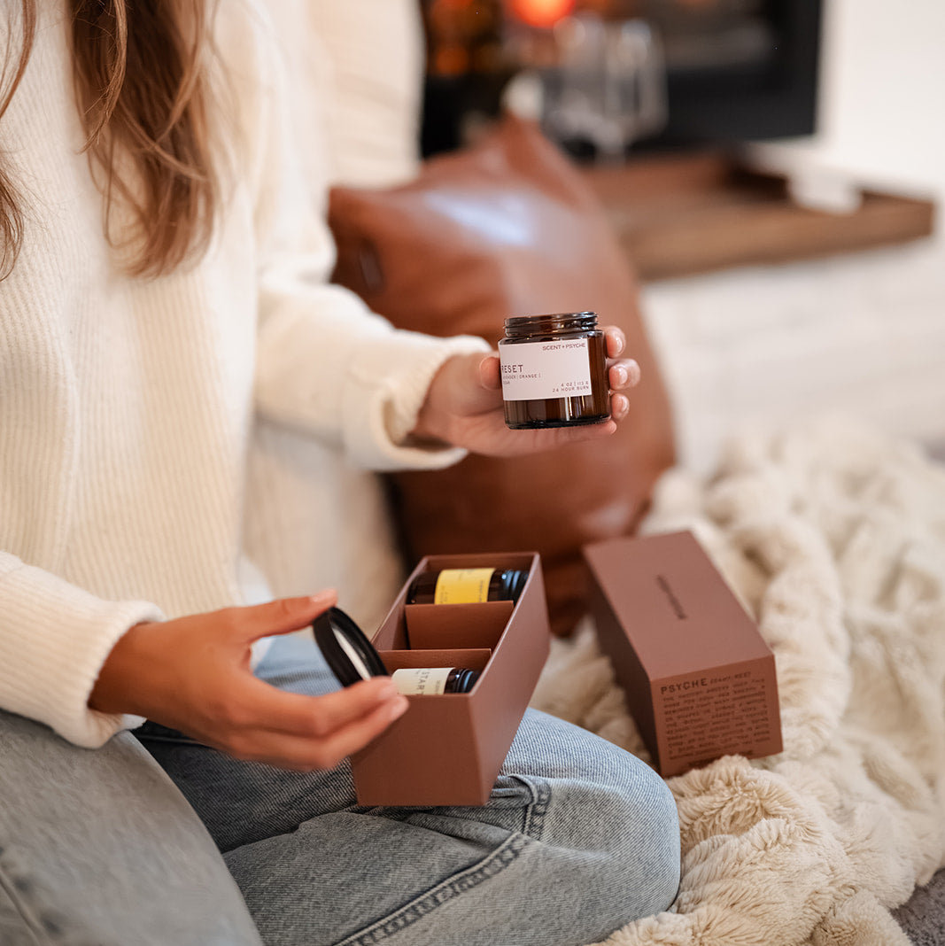 Scent + Psyche Energizing + Fresh soy candle discovery bundle in custom gift box. Scent + Psyche Energizing + Fresh soy candle discovery bundle in custom gift box. A woman sitting on a cozy rug in front of a fireplace, holding an amber glass wood wick candle and an open brown gift box with matching packaging.