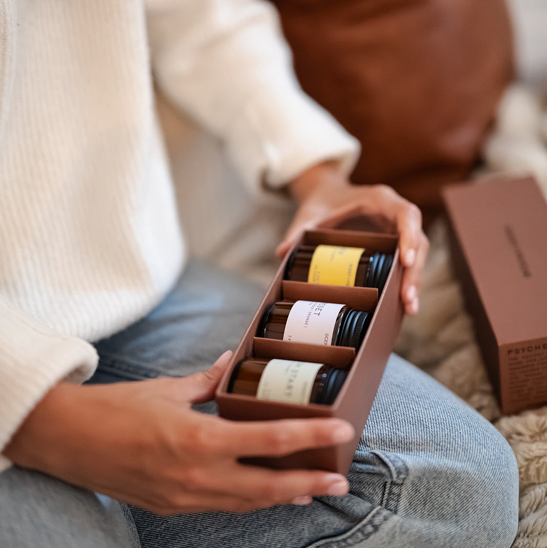 Scent + Psyche Energizing + Fresh soy candle discovery bundle in custom gift box. A person in a white sweater and blue jeans holds an open brown gift box containing  and wood wick candle gift set A matching brown box sits nearby on a textured rug, with a brown leather cushion in the background.